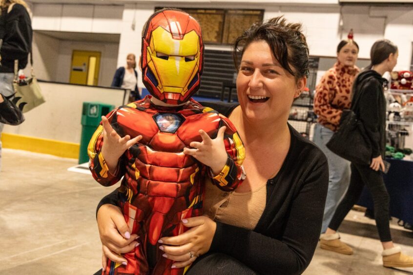 A boy dressed as Iron Man stands happily with his mum.