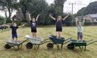 Four children standing in wheelbarrows with arms outstretched and cut grass behind them