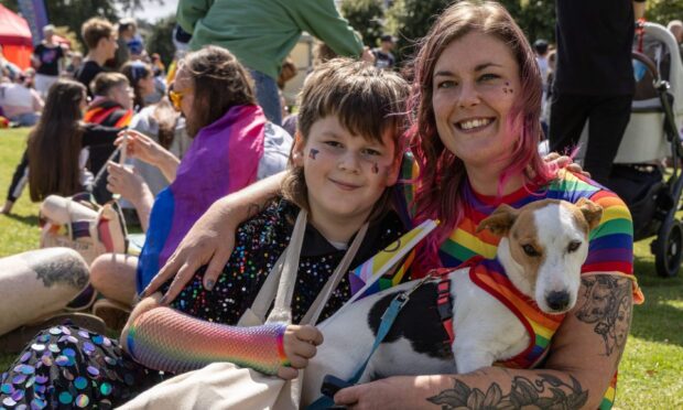 Two people smiling at Perthshire Pride, one wearing a rainbow-striped dress and holding a small white and brown dog, the other in a sequined black outfit with rainbow fishnet sleeves. Both have Pride face paint, and they are sitting on the grass among other attendees.