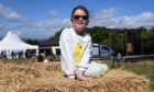 Smiling young girl wearing sunglasses and a lemon-print top sits on a hay bale at Kinross Show, with tents and visitors behind.