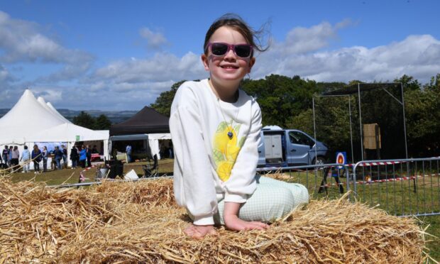 Smiling young girl wearing sunglasses and a lemon-print top sits on a hay bale at Kinross Show, with tents and visitors behind.