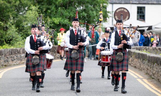 Doune Pipe Band in traditional dress playing bagpipes while marching over the Bridge of Dochart at Killin Highland Games 2025.