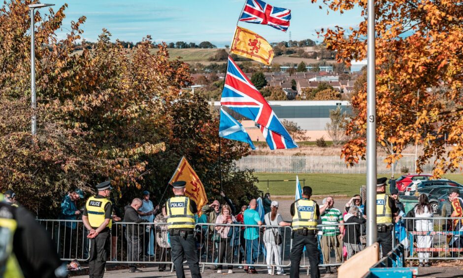 Anti-immigration protestors behind a barrier. Union Jack and Lion Rampant flags are held aloft. Police stand in the no-man's land between groups.