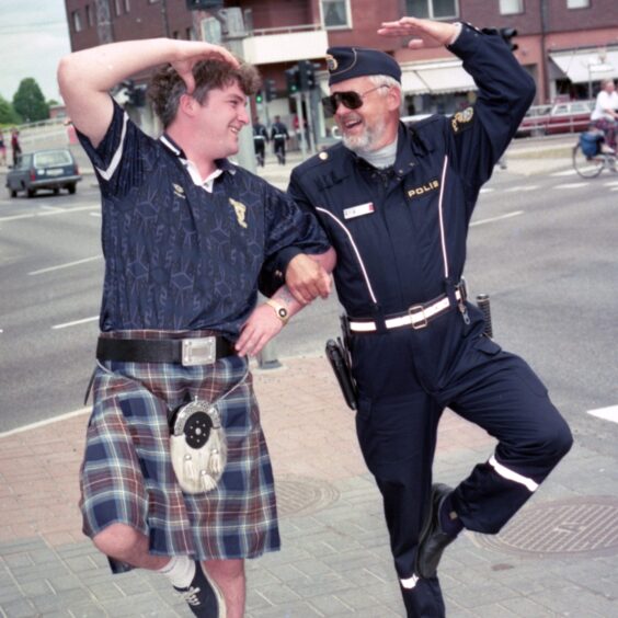 A Scotland fan wearing a replica top and kilt does the Highland fling with a Swedish policeman wearing a blue uniform and cap.
