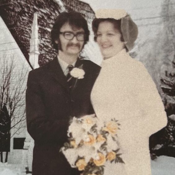 Erik Cramb on his wedding day wears glasses, a smart suit and tie and a corsage, while his wife has on a white dress and is holding a bouquet.