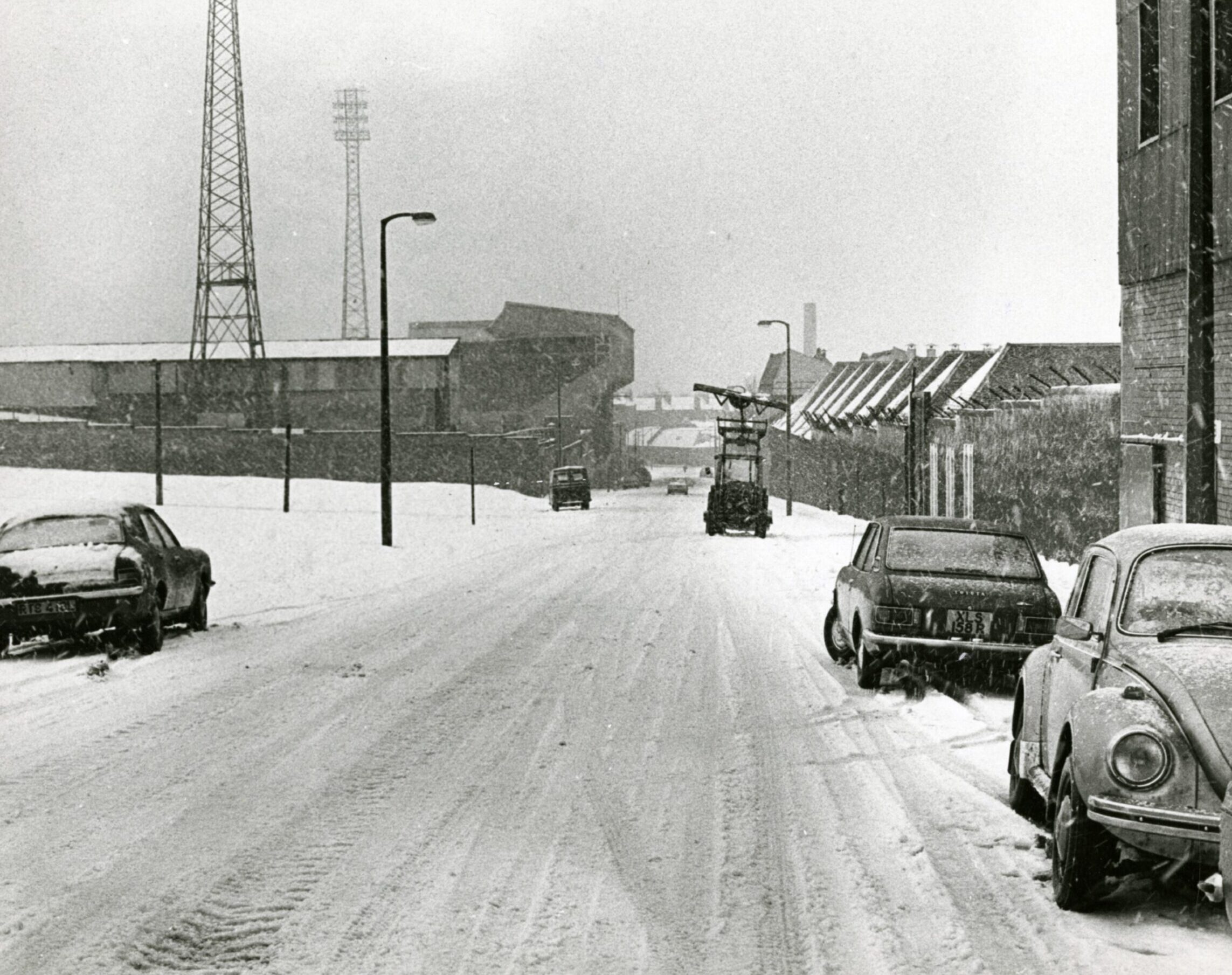 Snow covers the road looking down towards Tannadice Park. A few cars, including a VW Beetle, are parked at the side of the road and a tractor can be seen further down the street as snow continues to fall.