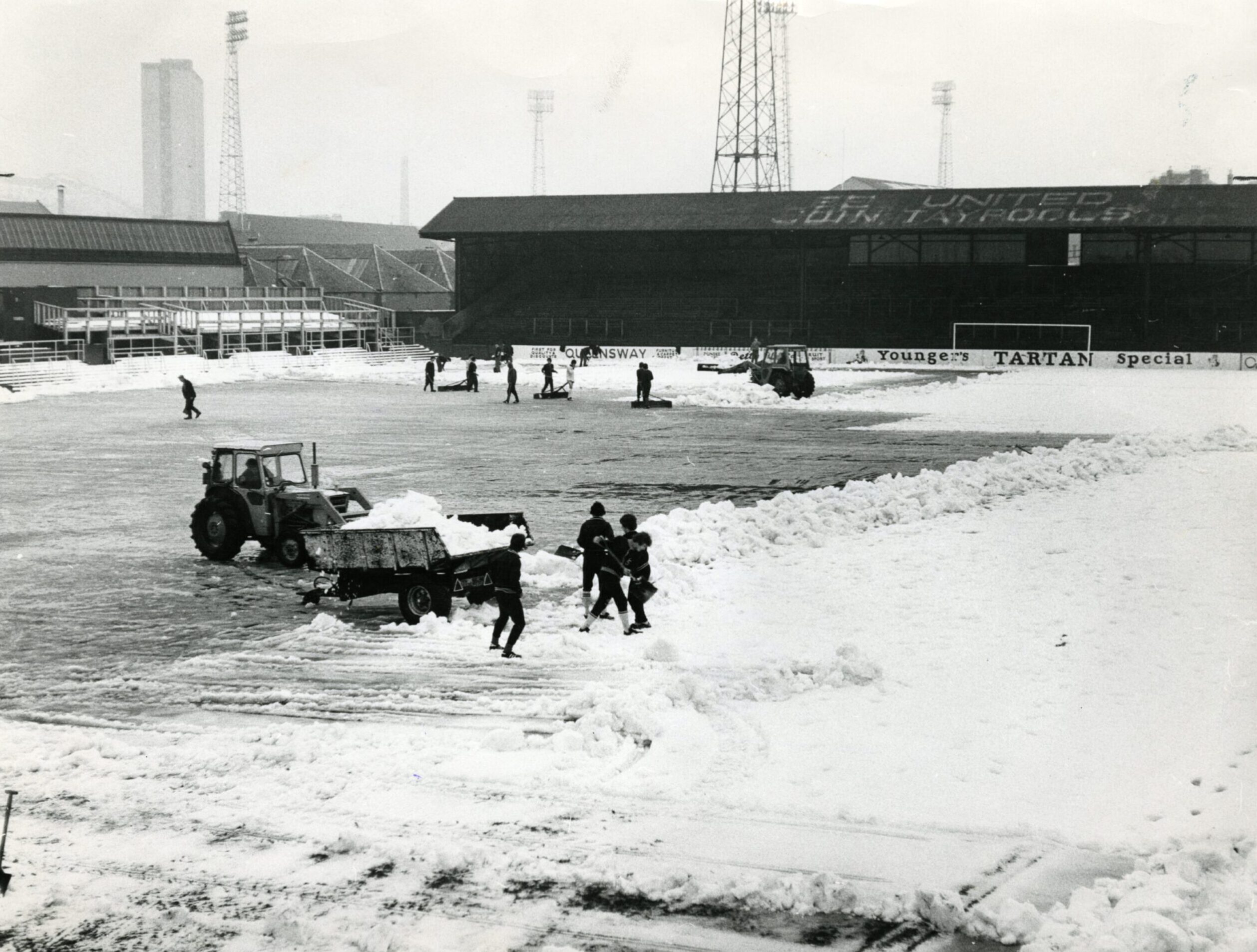 Players join in the effort to clear the pitch of thick snow. Two tractors have been brought in to help the operation and a bogey filled with snow is sitting on the pitch,
