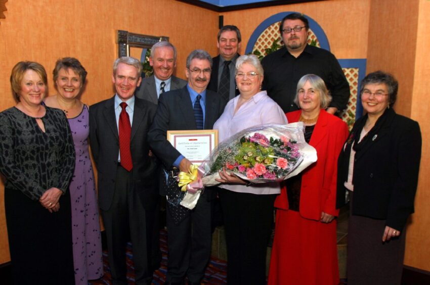 Erik Cramb and wife Elizabeth with well-wishers at a retiral event. Erik is holing a certificate and Elizabeth has a bunch of flowers.