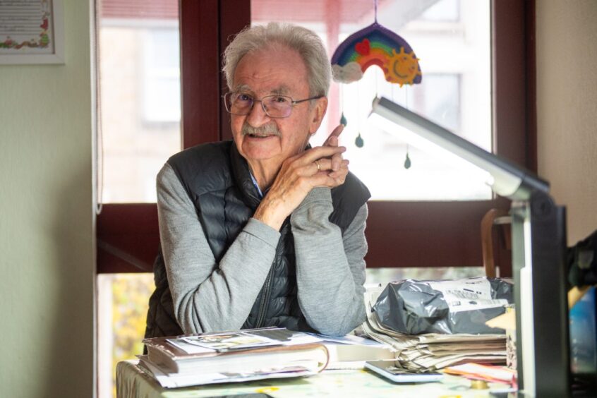 A man wearing glasses, a grey jumper and black gilet stands at a desk covered with books and cuttings.