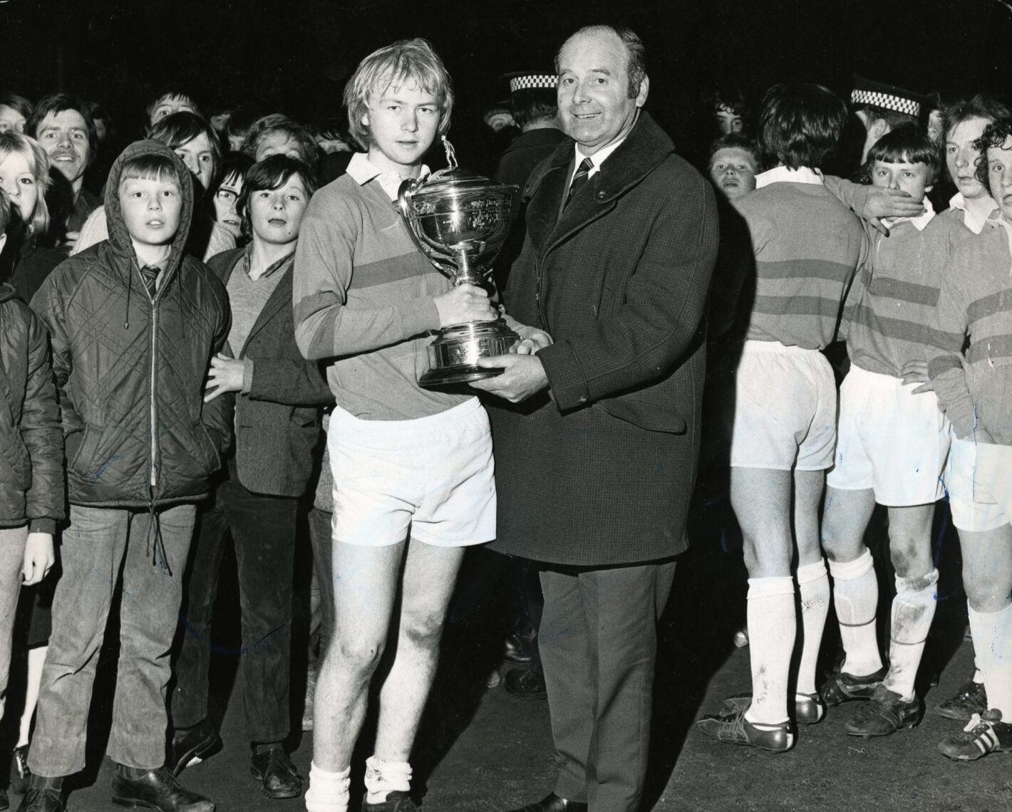 Dundee United manager Jerry Kerr hands over a cup to blond-haired Kirkton High School captain Paul Tovey. Other members of the team and young pupils are in the background.