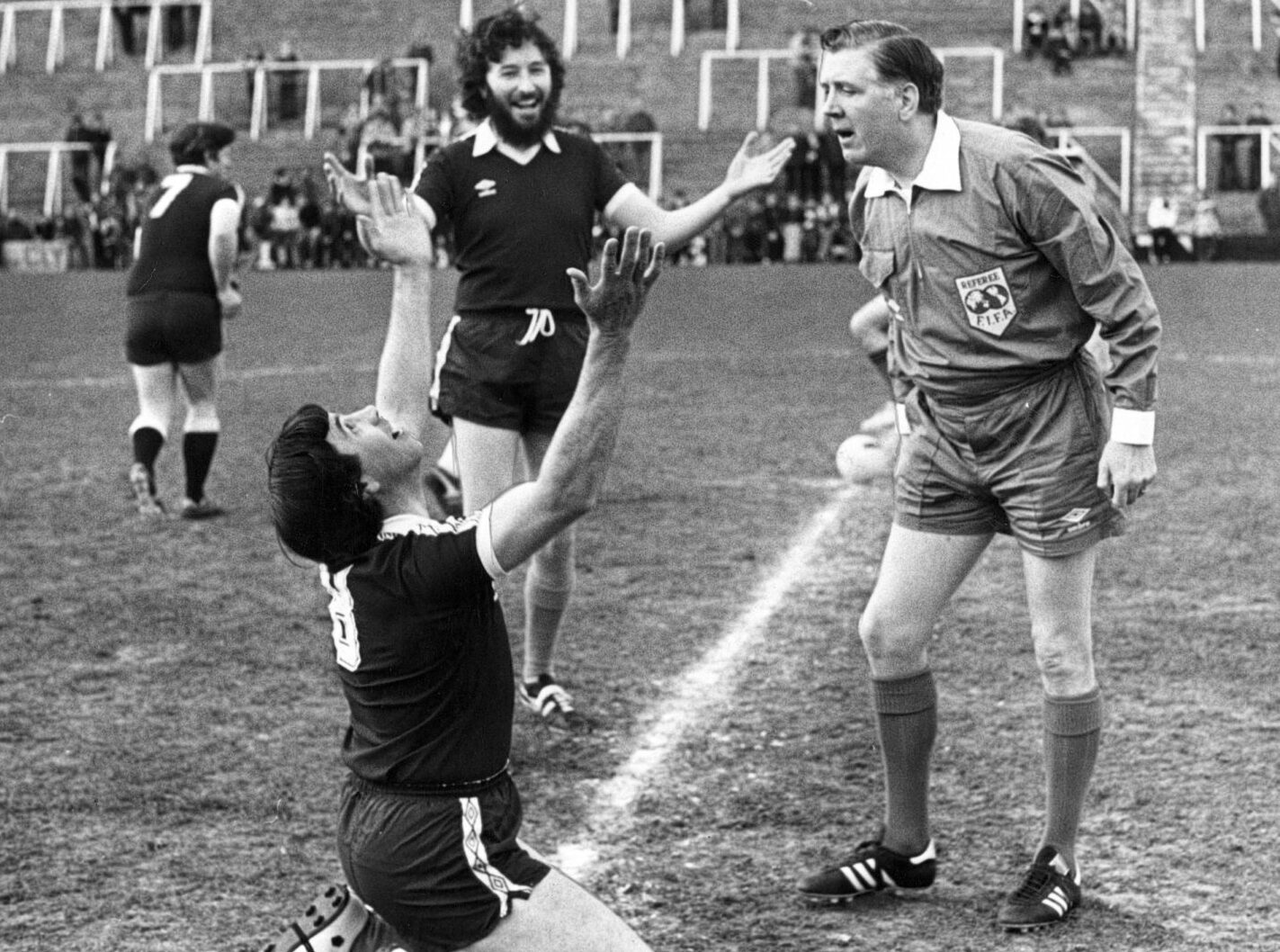 Players taking part in a charity game between ministers ands butchers pretend to seek divine intervention in front of the referee. One player is kneeling on the pitch while raising his arms to the heavens as the ref looks over him.