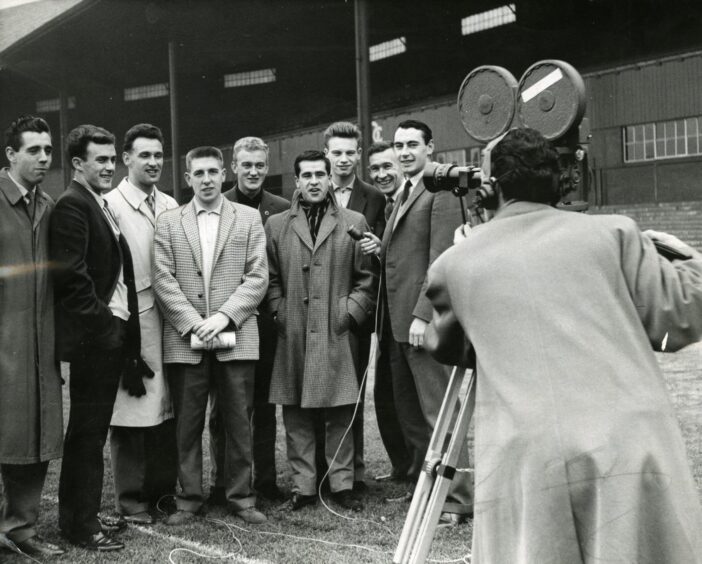 a group of players stands on the pitch while a man operates a camera for a television interview at Dens in 1961