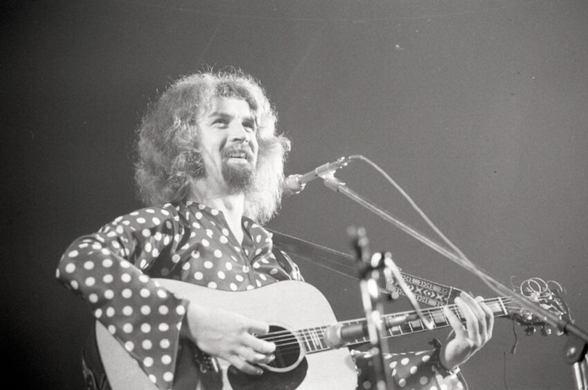 a smiling Connolly wearing a polka dot shirt on stage at the Caird Hall. 