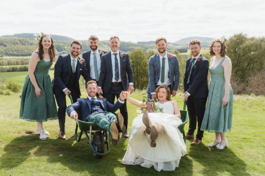 Bride and groom sitting in wheelbarrows with their friends in the background.