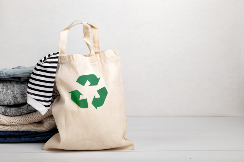 A canvas tote bag featuring the recycling logo, sitting next to a pile of clothes.