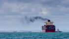 Black smoke pours out of a cargo container ship as it pulls into port.