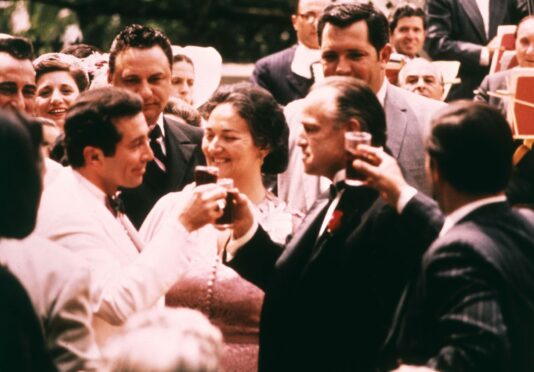 Don Vito Corleone (played by Marlon Brando), right, clinks glasses with wedding guest Johnny Fontane (Al Martino) as wife Carmela (Morgana King) stands by and smiles in The Godfather