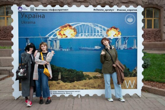 Young women in Kyiv pose in front of a giant stamp hailing the Kerch bridge explosion last week