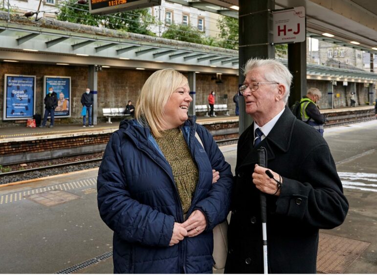 Man and woman on train platform. Smiling and laughing. Man carrying long cane.