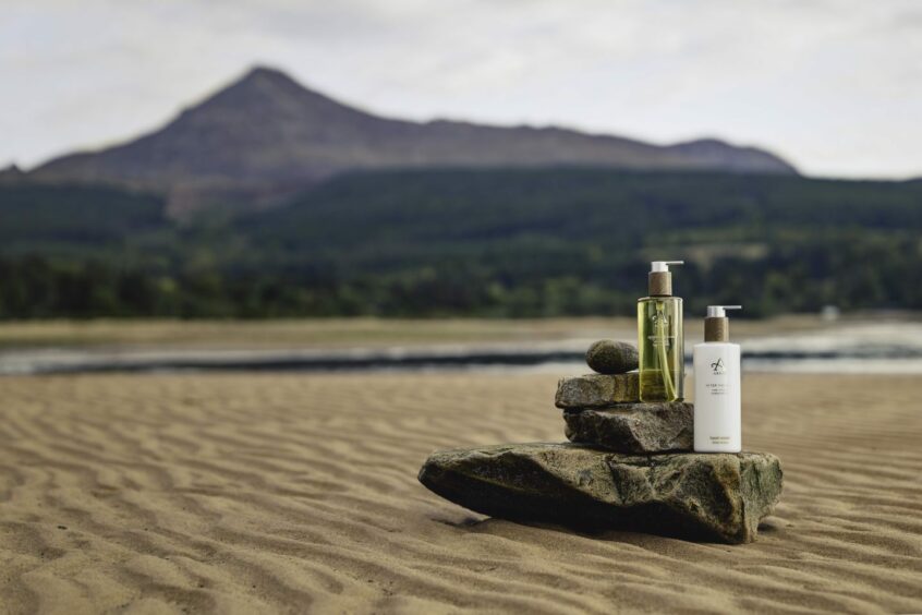 bottles of Arran Sense of Scotland's bath, body and home fragrance gifts set against the backdrop of a beach, one of the places to visit in the west coast of Scotland