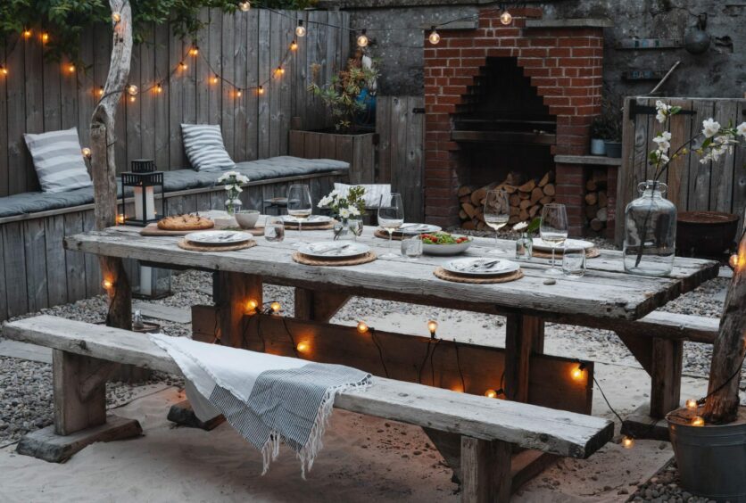 A wooden table and two matching benches are set for dinner in a cosy and well-lit setting with an outdoor brick fireplace.