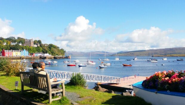 Couple sat on bend admiring a beautiful view of a Scottish harbour in the sun.