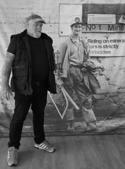 Donald Fulton from Muirkirk, East Ayrshire, with his lamp, in front of a banner displaying a Milton Rogovin photograph from 1982.