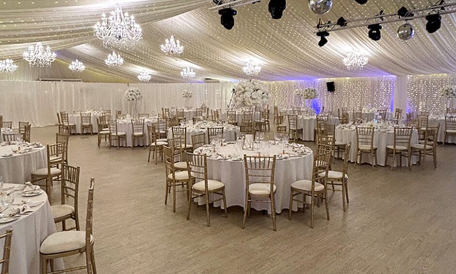 wedding reception setup inside a marquee with lit chandeliers, round tables, chairs and table settings 