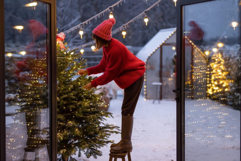 woman stands on top of a stool to decorate an outdoor Christmas tree