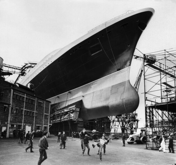 Workers play football beneath the QE2’s massive hull.