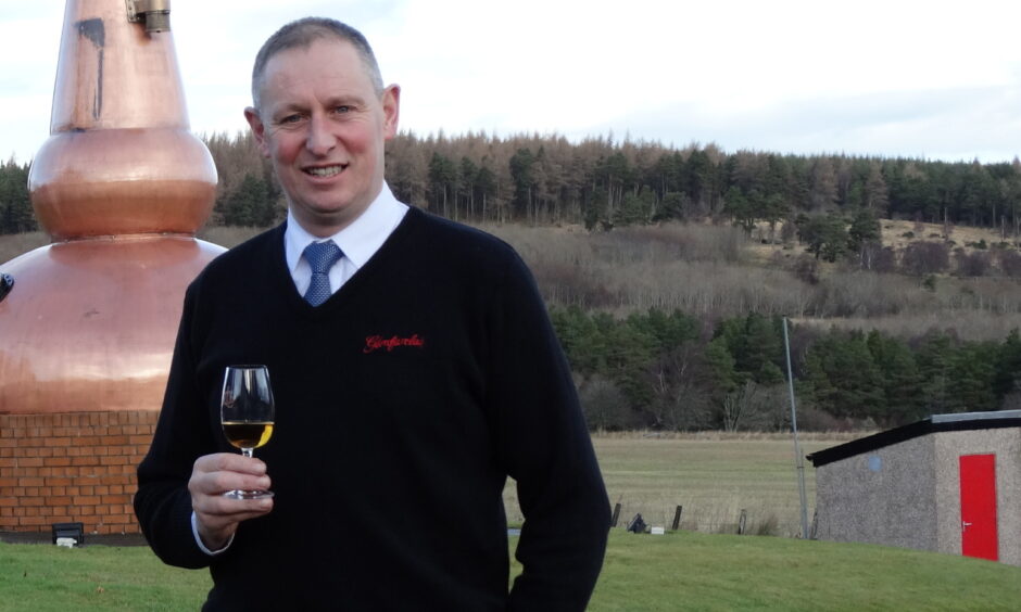 Picture of Callum Fraser the manager of the Glenfarclas Distillery. He is wearing a black jumper on top of a shirt and tie and is clean shaven. He is holding a glass of whisky. 