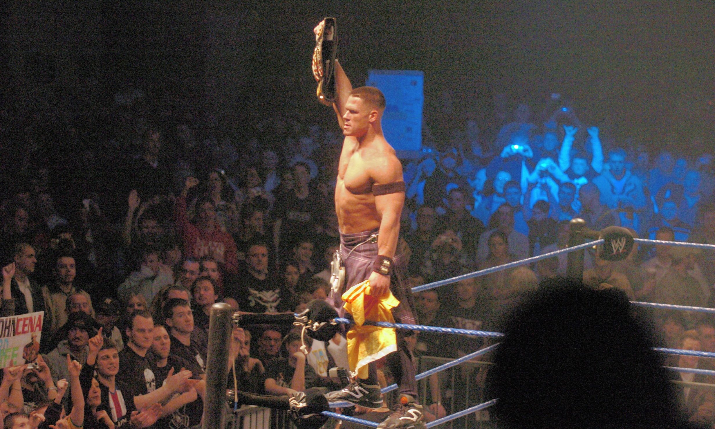 John Cena wearing a kilt and standing on the ropes of the ring, holding a belt above his head