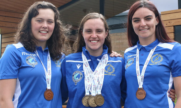 Three female swimmers stand next to each other, smiling, wearing medals around their necks.