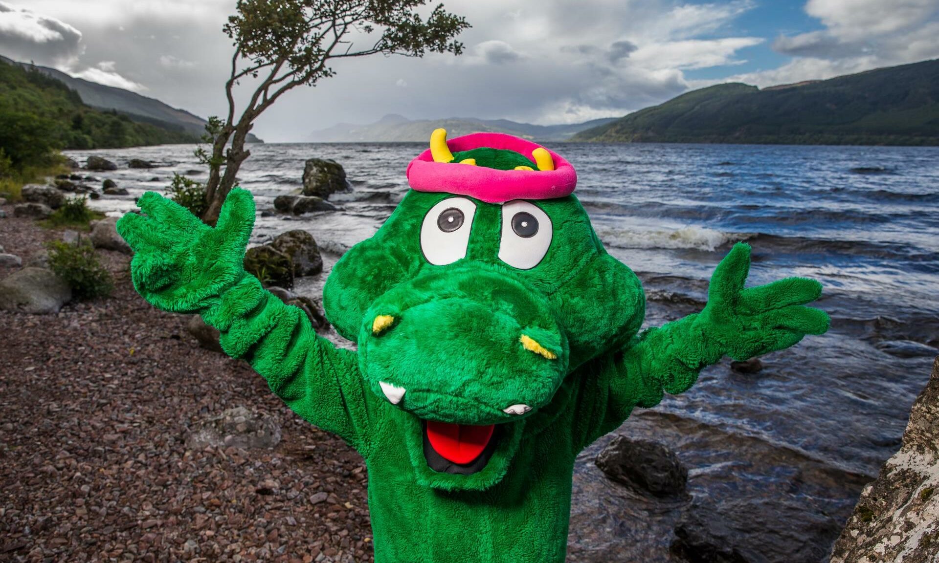 A Loch Ness Monster mascot standing in front of Loch Ness