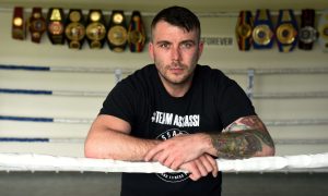 Aberdeen boxing legend Lee McAlliste leans over the ropes of a boxing ring with his title belts on display behind him.