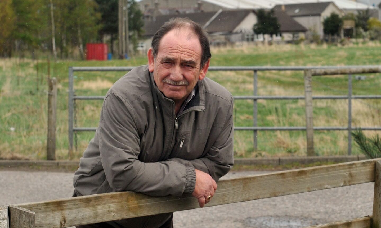 Alastair Kennedy leaning on a fence at the old mart in Elgin with grass behind him. 