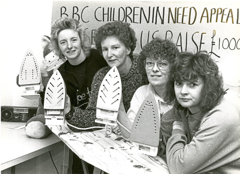 Four women holding irons and leaning on an ironing board, behind them is a sign reading "BB Children In Need appeal"