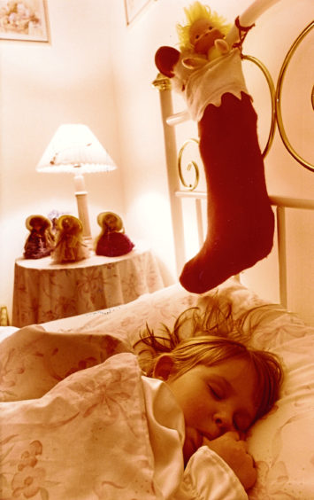 A child sleeps soundly before Christmas, while a stocking filled with toys hangs on their bed