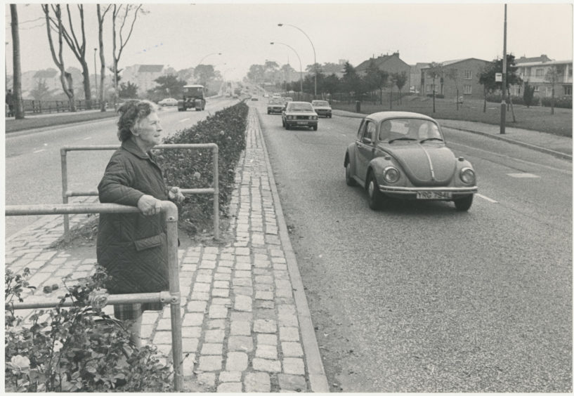 Regular crossing user Mrs Margaret Tosh of Woodhill Court, Aberdeen, steadies herself with a hand rail as she waits for a clear stretch to cross Anderson Drive for her bus.  12 October 1979.