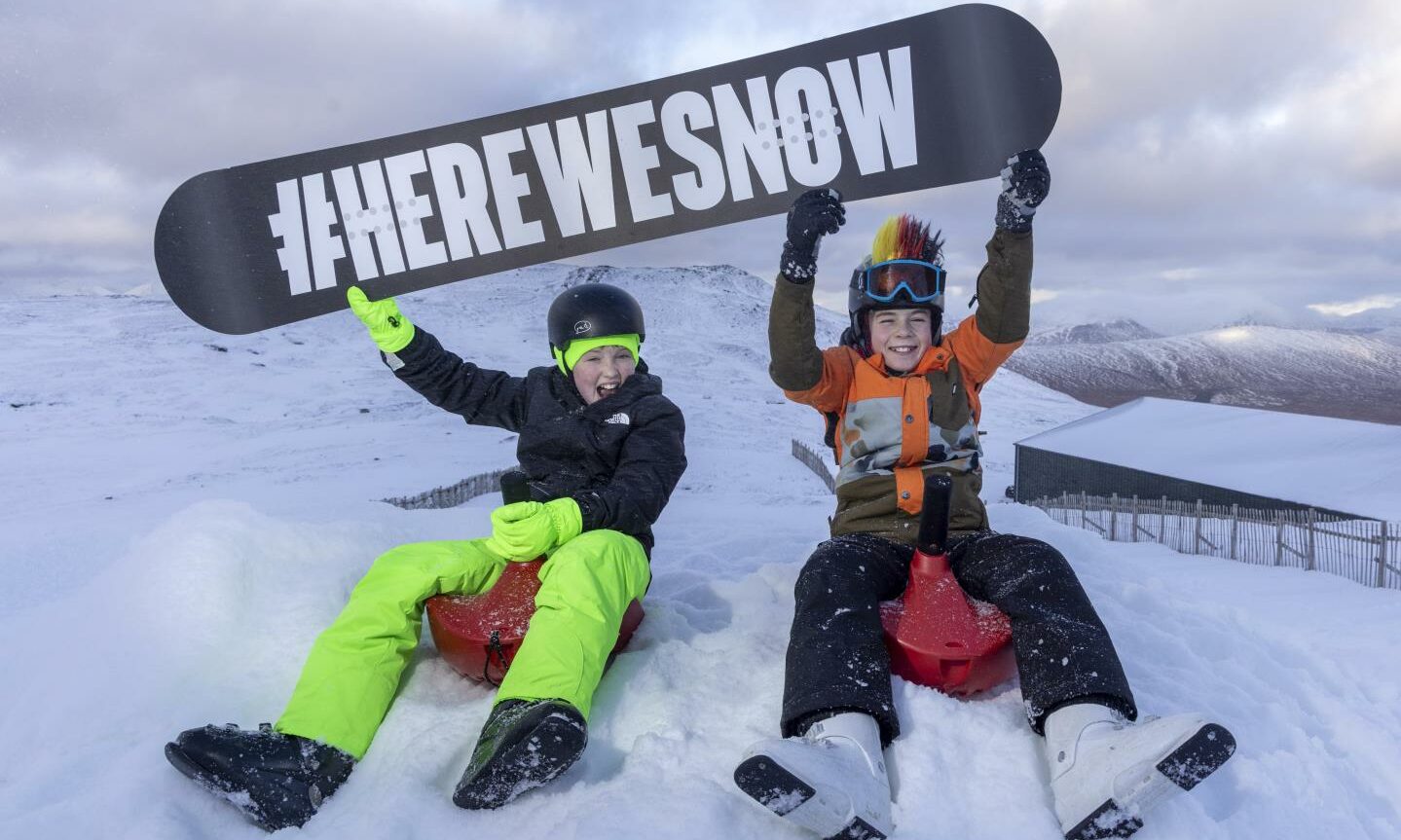 Ben Joshua and Kody Cumming at Glencoe ski centre holding snowboard saying Here We Snow.