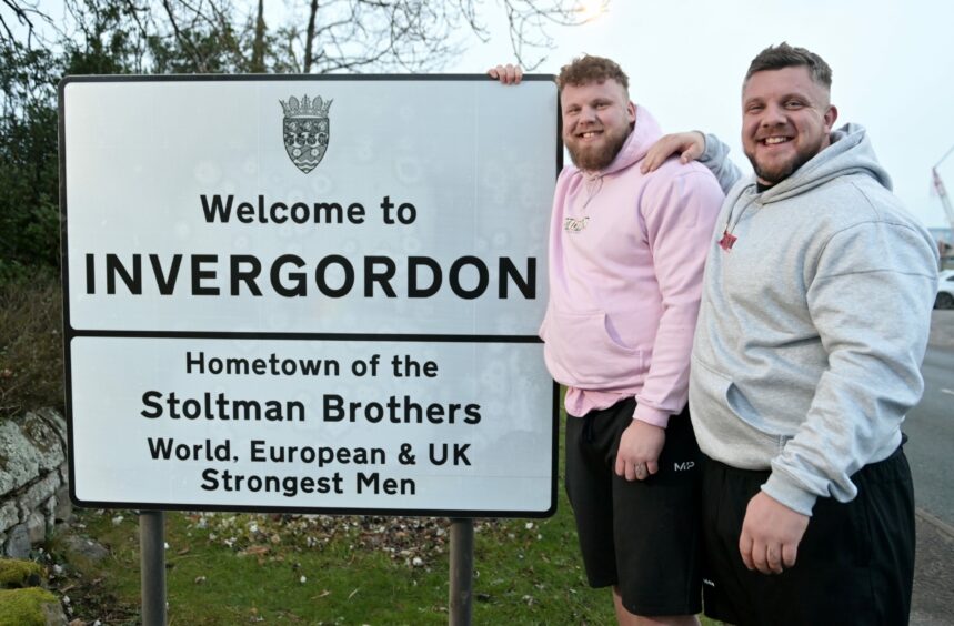 Stoltman brothers in front of Invergordon welcome sign with text: "Welcome to Invergordon. Hometown of the Stoltman Brothers. World, European & UK Strongest Men"