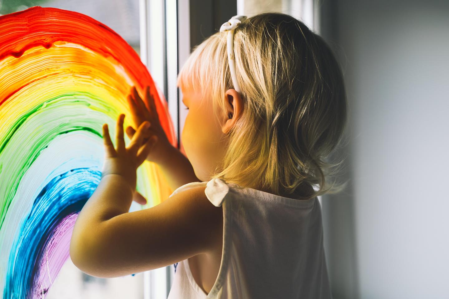 A child indoors with a rainbow painted on a window