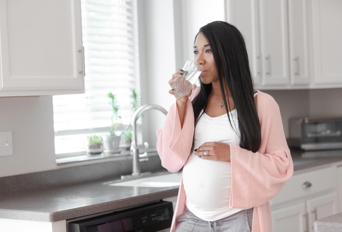 Pregnant woman drinking a glass of water by the sink 