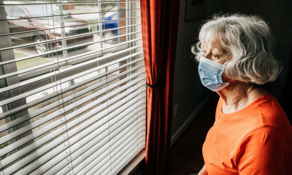 Elderly woman wearing a protective face mask looking out of the window with blinds. 
