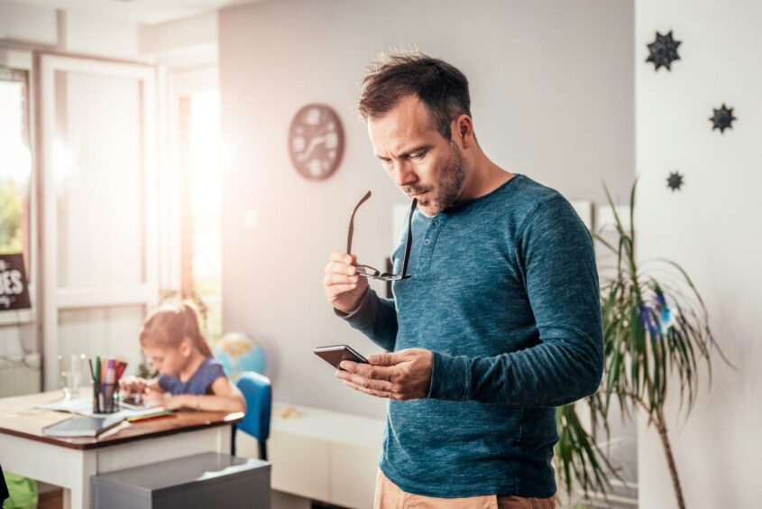 Father texting on his phone whilst his daughter is in the background sitting at the table doing her homework