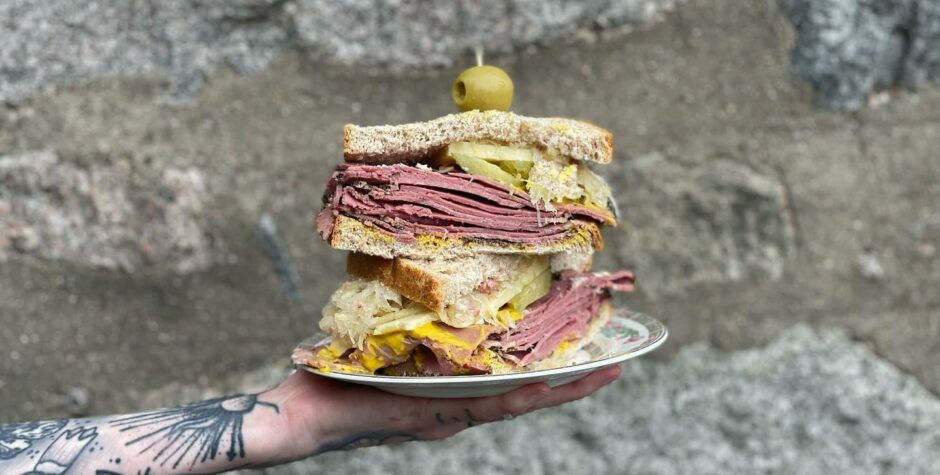 A hand holding a plate with a meaty deli sandwich 