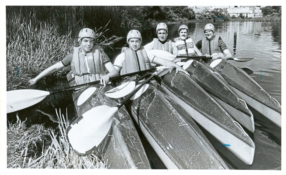 Black and white photo of five men in helmets and life jackets posing in canoes
