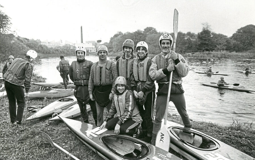 A group of canoeists from Aberdeen Kayak Club with helmets and life jackets pose by two canoes on the shore