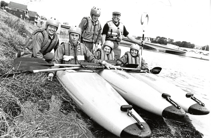 Six canoeists in helmets and life jackets pose for a photo on the shore. Three sit in canoes and three stand behind