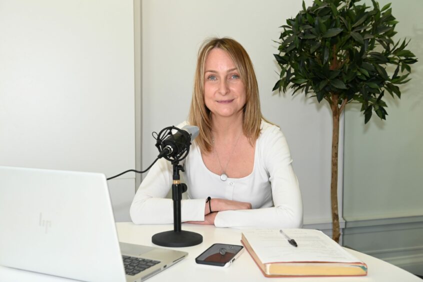 Isla Stewart sitting in front of a black microphone at a white table with a laptop, a phone, a book, pen and paper in front of her